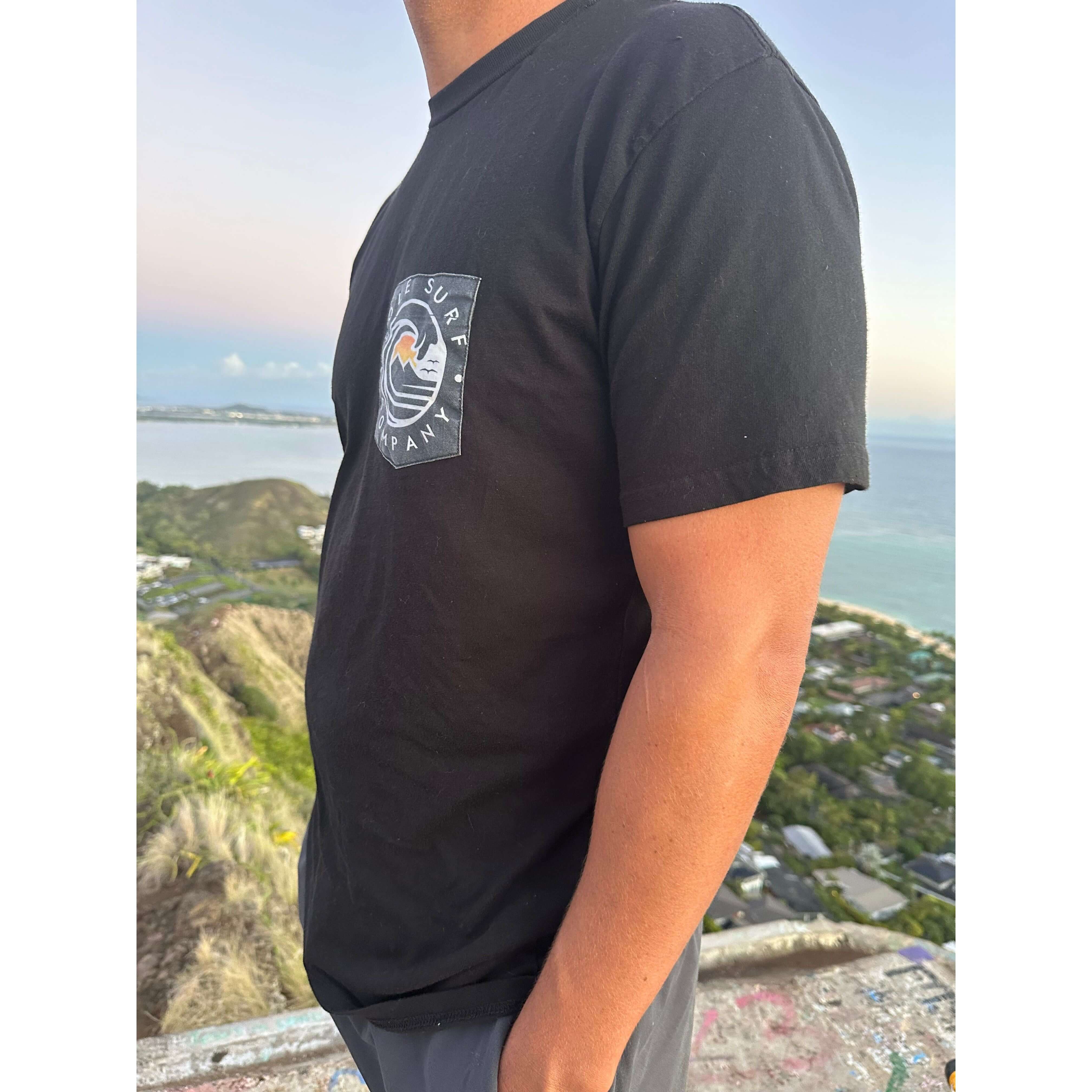 Black logo pocket tee worn outdoors, perfect for casual beachwear, made of soft premium cotton.