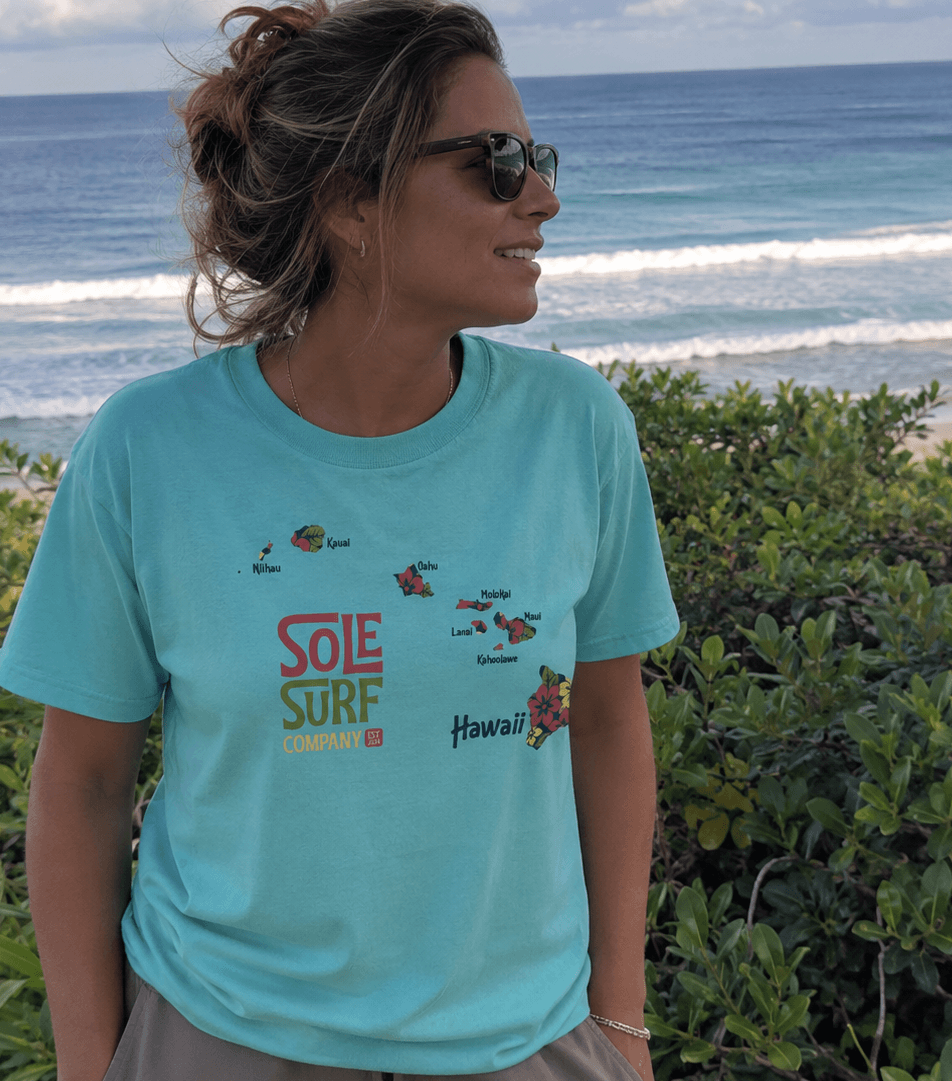 A woman wearing a light blue graphic t-shirt with 'SOLE SURF COMPANY' and 'Hawaii' printed on the front, standing outdoors on a beach with the ocean and green foliage in the background.