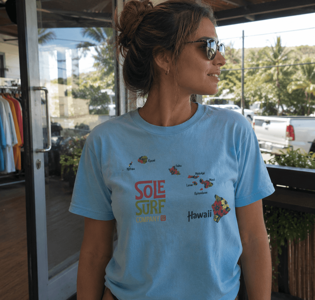 A woman wearing a light blue graphic t-shirt with 'Sole Surf Company' and 'Hawaii' printed on the front, standing on a covered outdoor walkway.