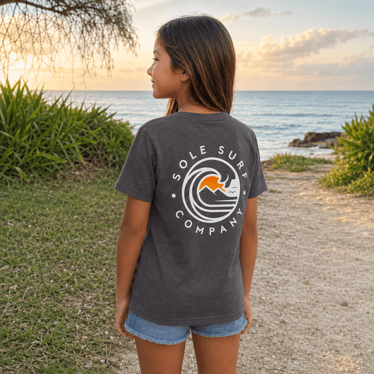A young girl standing on a beach path at sunset, wearing a dark gray t-shirt with a white 'Sole Surf Company' wave graphic on the back and denim shorts.