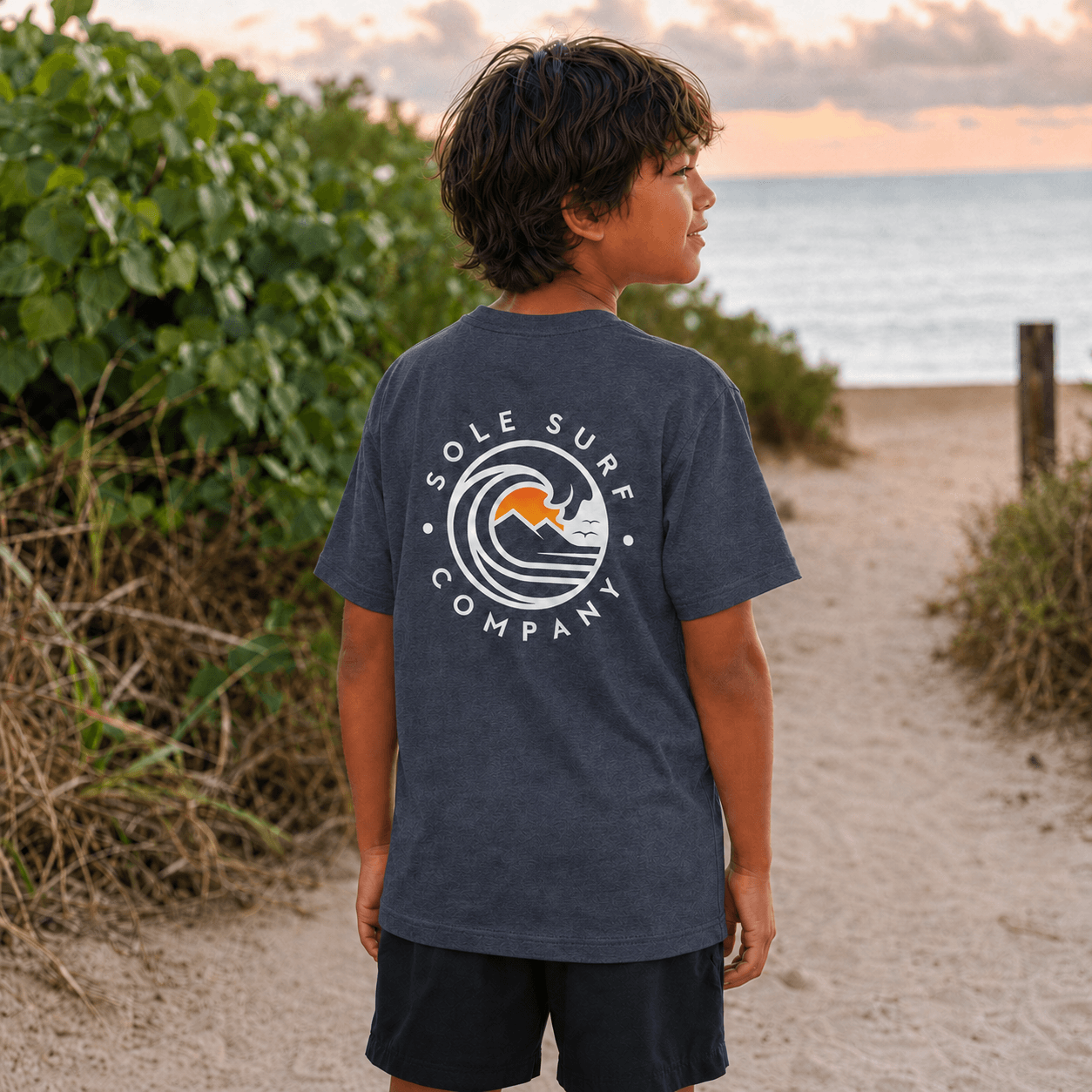 A child wearing a dark heather gray t-shirt with a white and orange 'Sole Surf Company' wave graphic on the back, standing on a sandy beach path with the ocean and green foliage in the background.