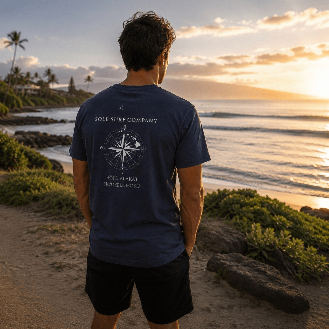 Person wearing a blue t-shirt with a compass design, standing on a beach path at sunset.