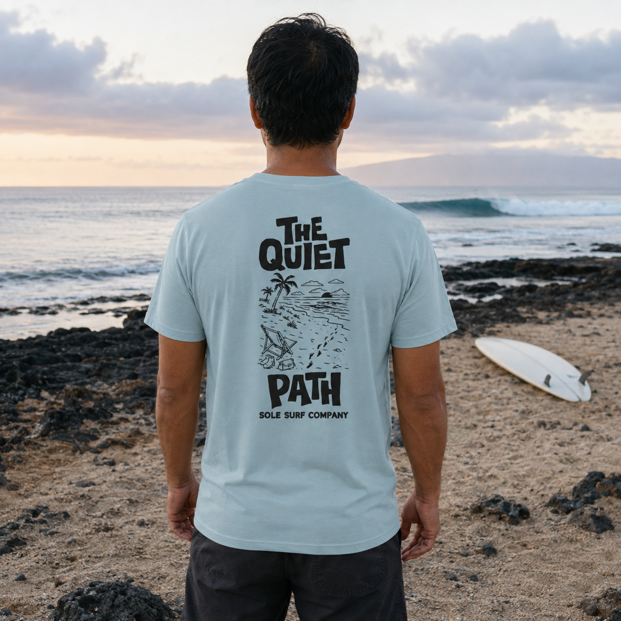 A man stands on a beach facing the ocean, wearing a light blue t-shirt with a black graphic print on the back that reads 'THE QUIET PATH SOLE SURF COMPANY'. A white surfboard lies on the sand in the background near the water.