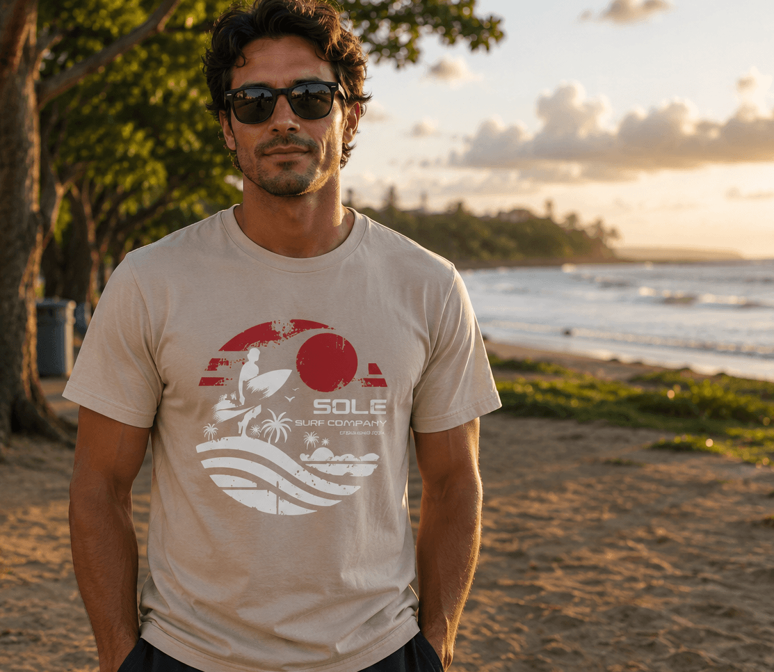 Man wearing a beige t-shirt with a graphic design standing on a beach at sunset.