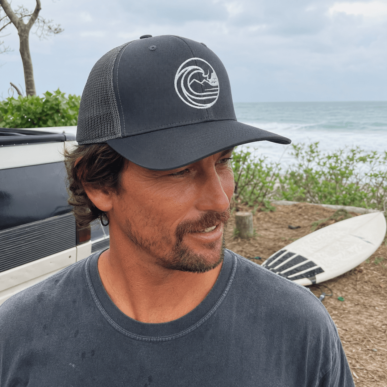A man wearing a dark gray mesh-back trucker hat with a white wave logo and a dark gray t-shirt, standing outdoors near the ocean with a surfboard and a vehicle in the background.