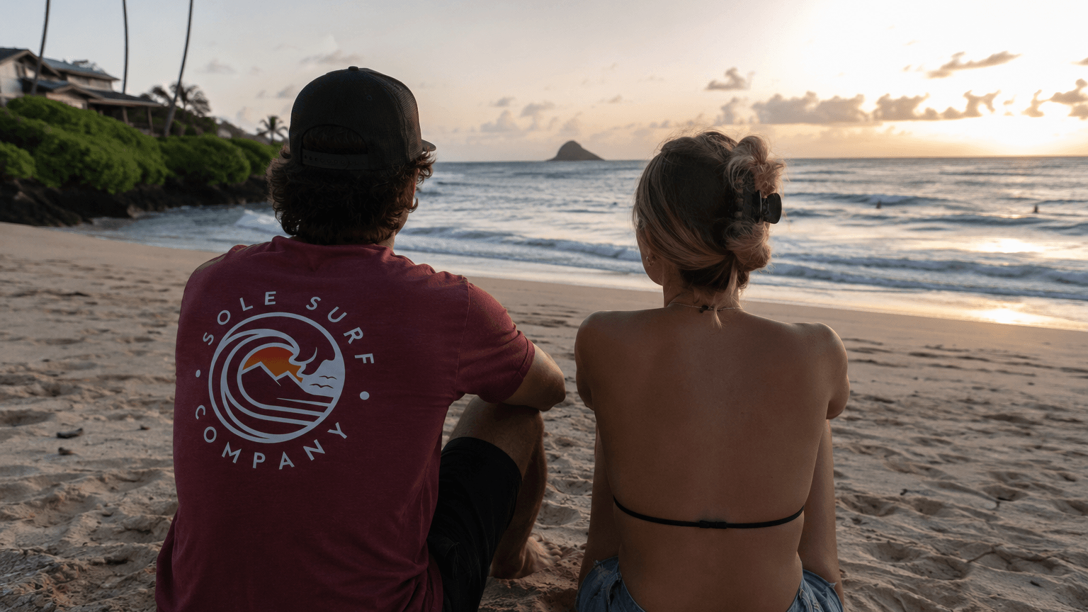 Two people sitting on a beach at sunset, one wearing a 'Sole Surf Company' t-shirt.