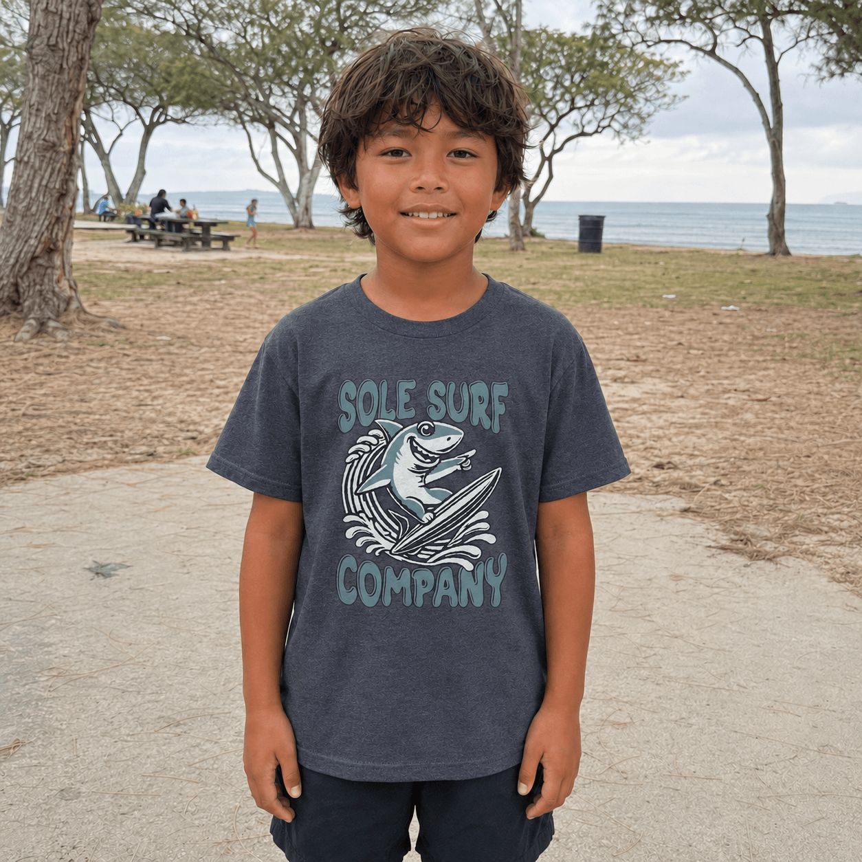 A young boy stands on a beach path wearing a dark gray t-shirt with a light blue graphic that reads 'SOLE SURF COMPANY' and shows a shark on a surfboard.