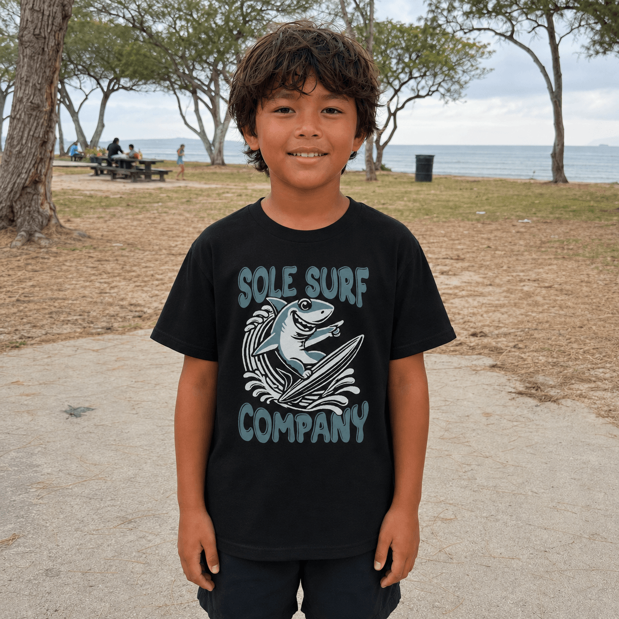 A young boy stands outdoors in a park near the beach wearing a black graphic t-shirt with a shark and wave design and the text 'SOLE SURF COMPANY'.