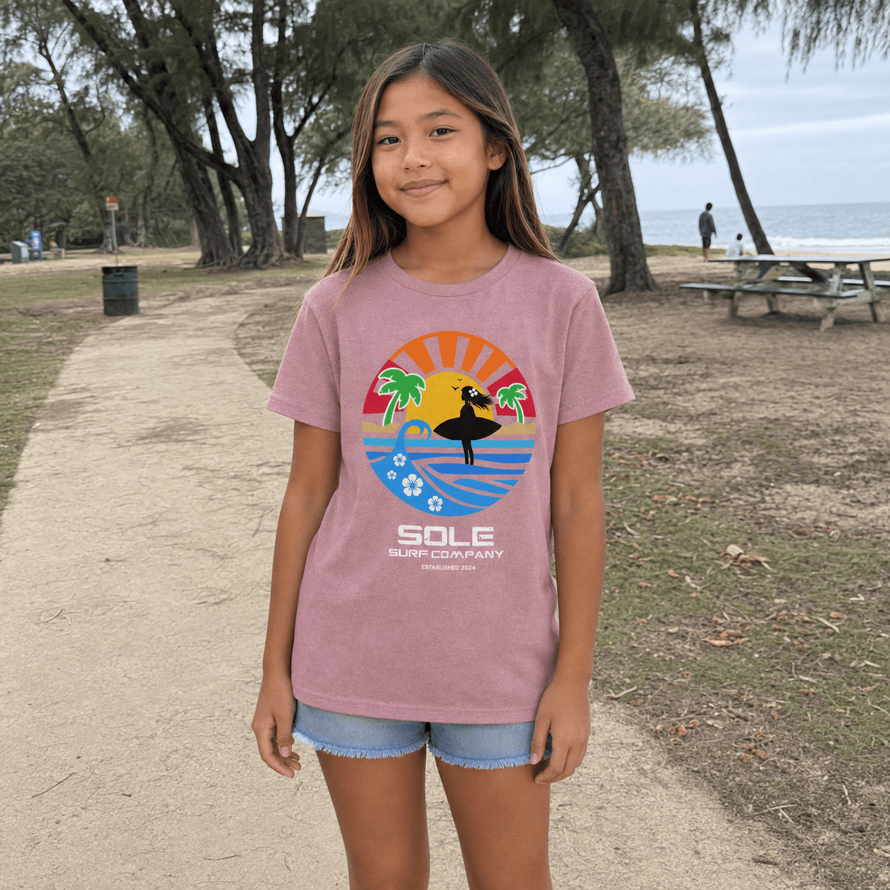 A young girl stands on a beach path wearing a dusty pink t-shirt with a colorful graphic of a surfer and the text 'SOLE SURF COMPANY'.