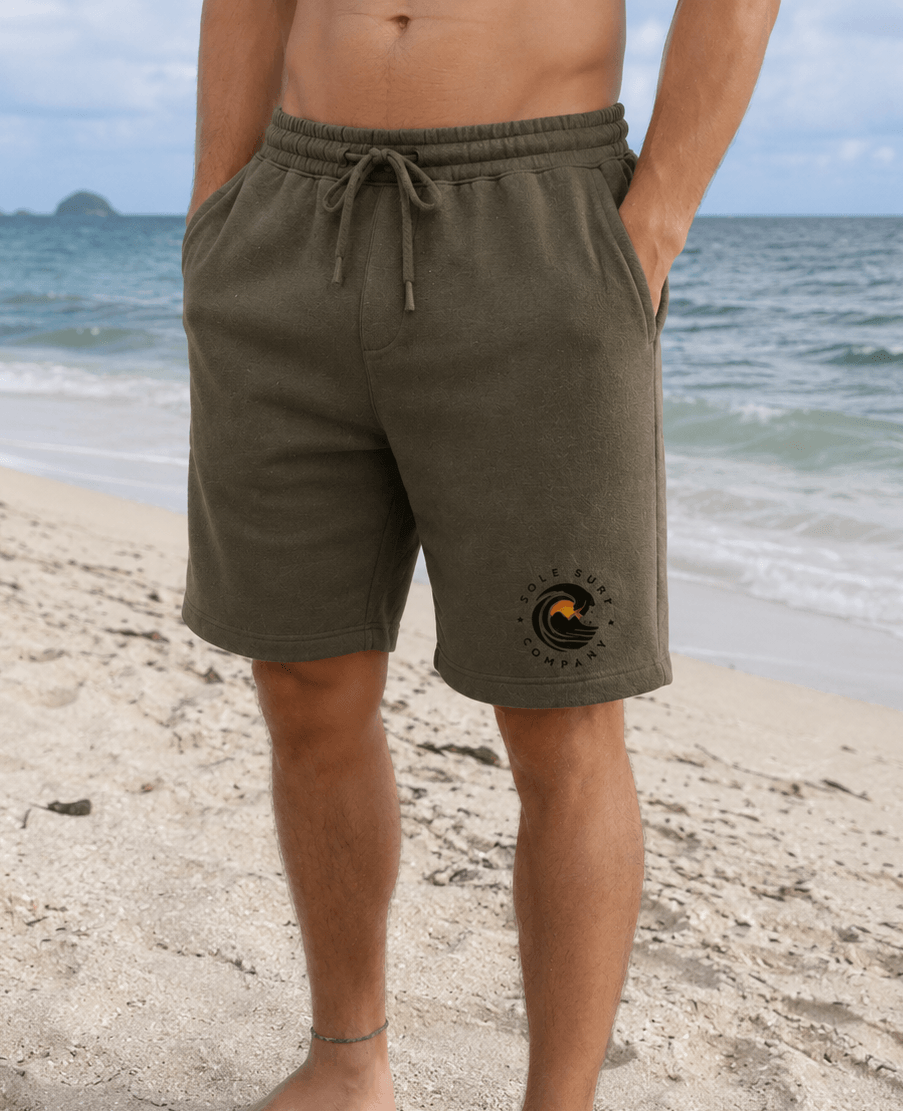 A man standing on a sandy beach wearing olive green drawstring shorts with a circular black and yellow logo on the lower left leg. The background shows the ocean and cloudy sky.