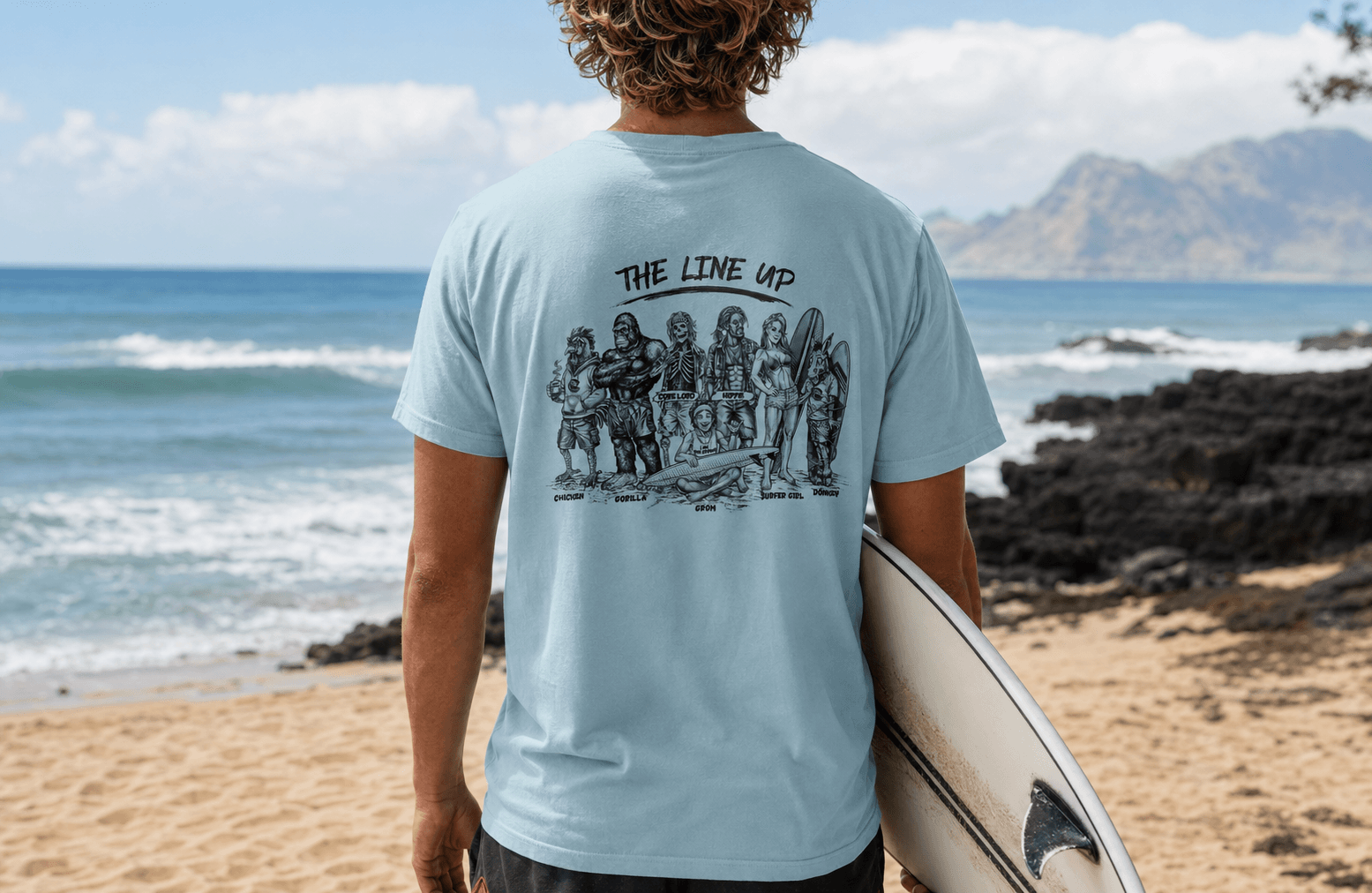 A man standing on a beach, viewed from behind, wearing a light blue t-shirt with a black graphic print of surfers and the text 'THE LINE UP'. He is holding a white surfboard and looking out towards the ocean and mountains.
