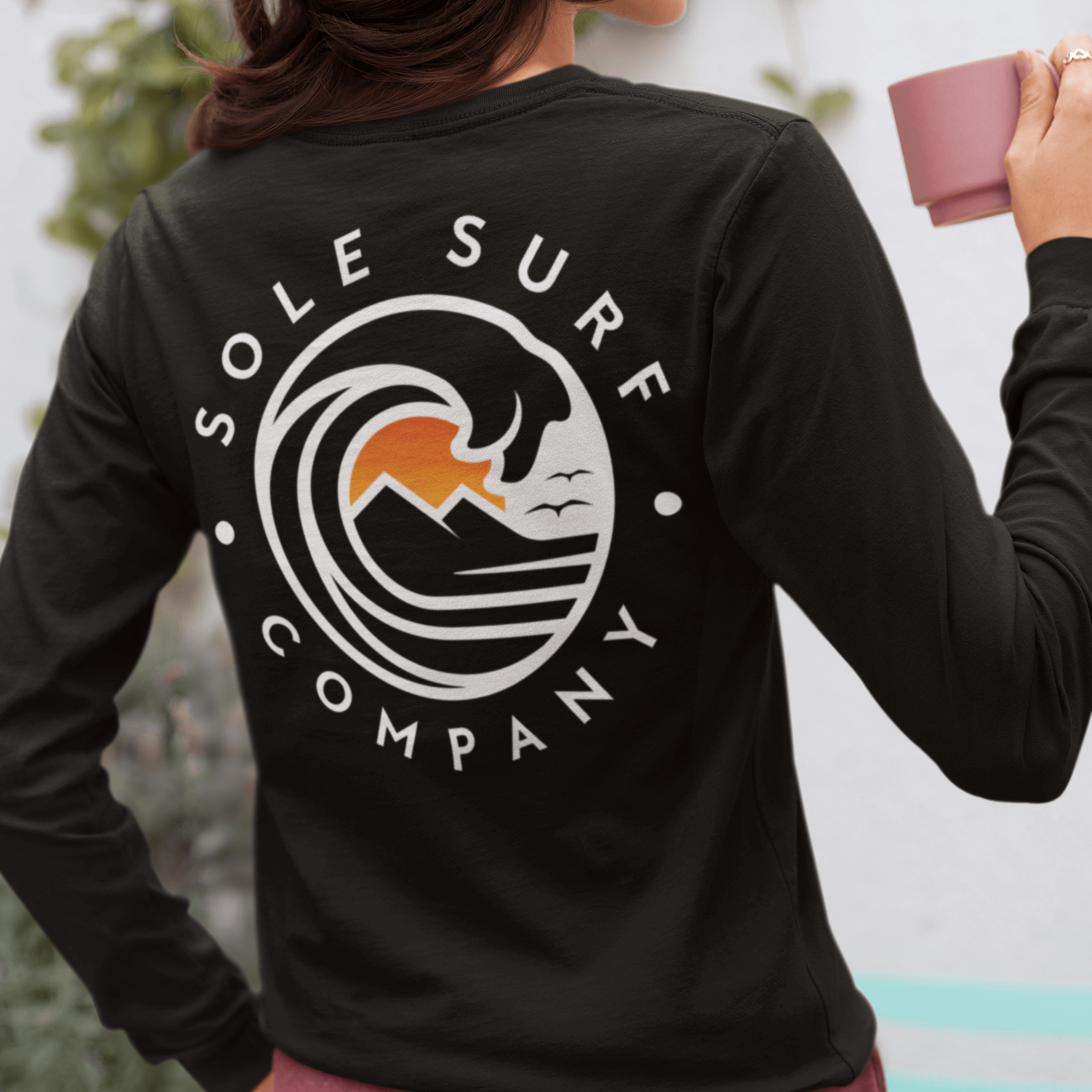 A woman wearing an SSC White Logo Long Sleeve surf tee, enjoying a drink with a coastal-themed design on the back.