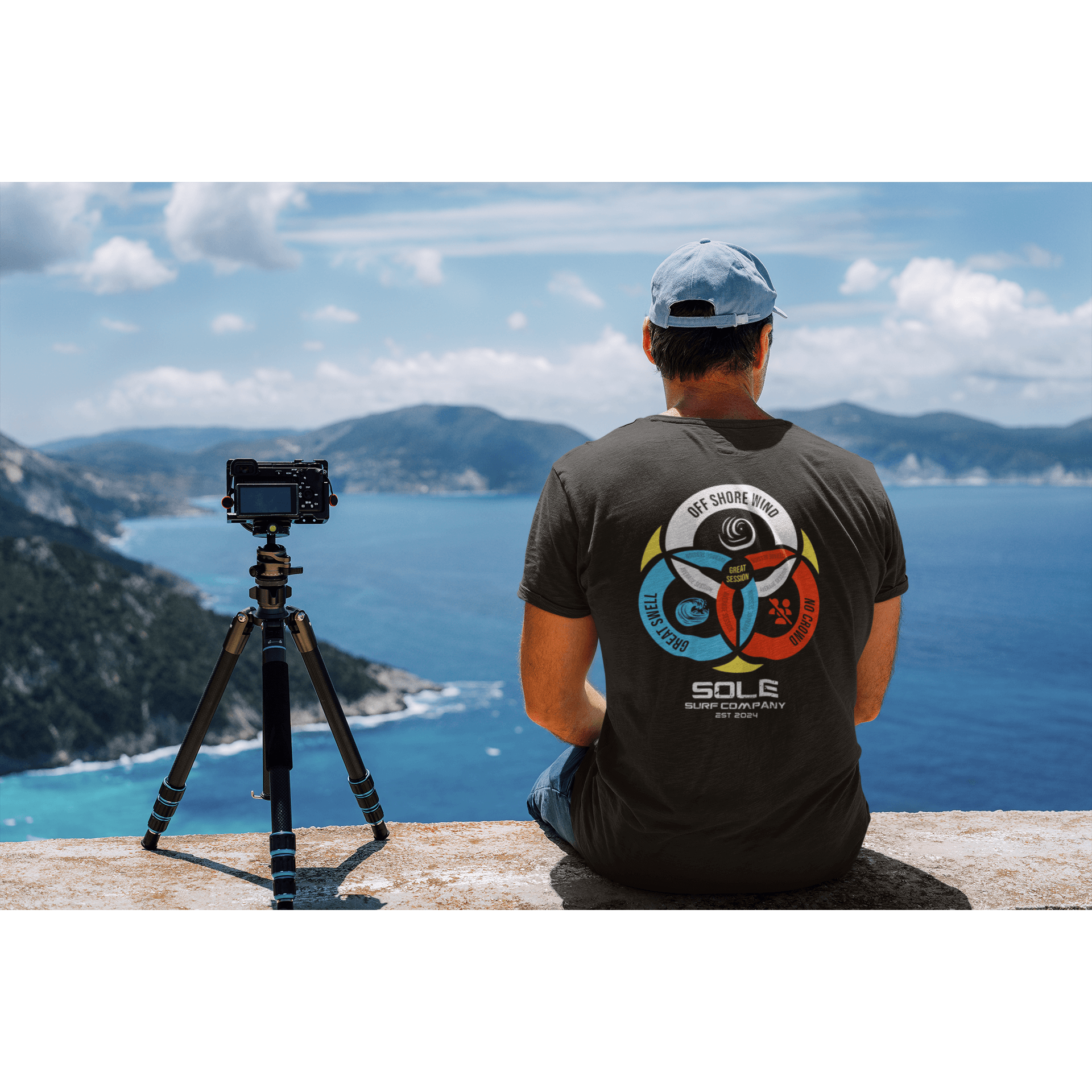 Man wearing a T-shirt with surf-inspired design, capturing scenic coastal views with camera on tripod.