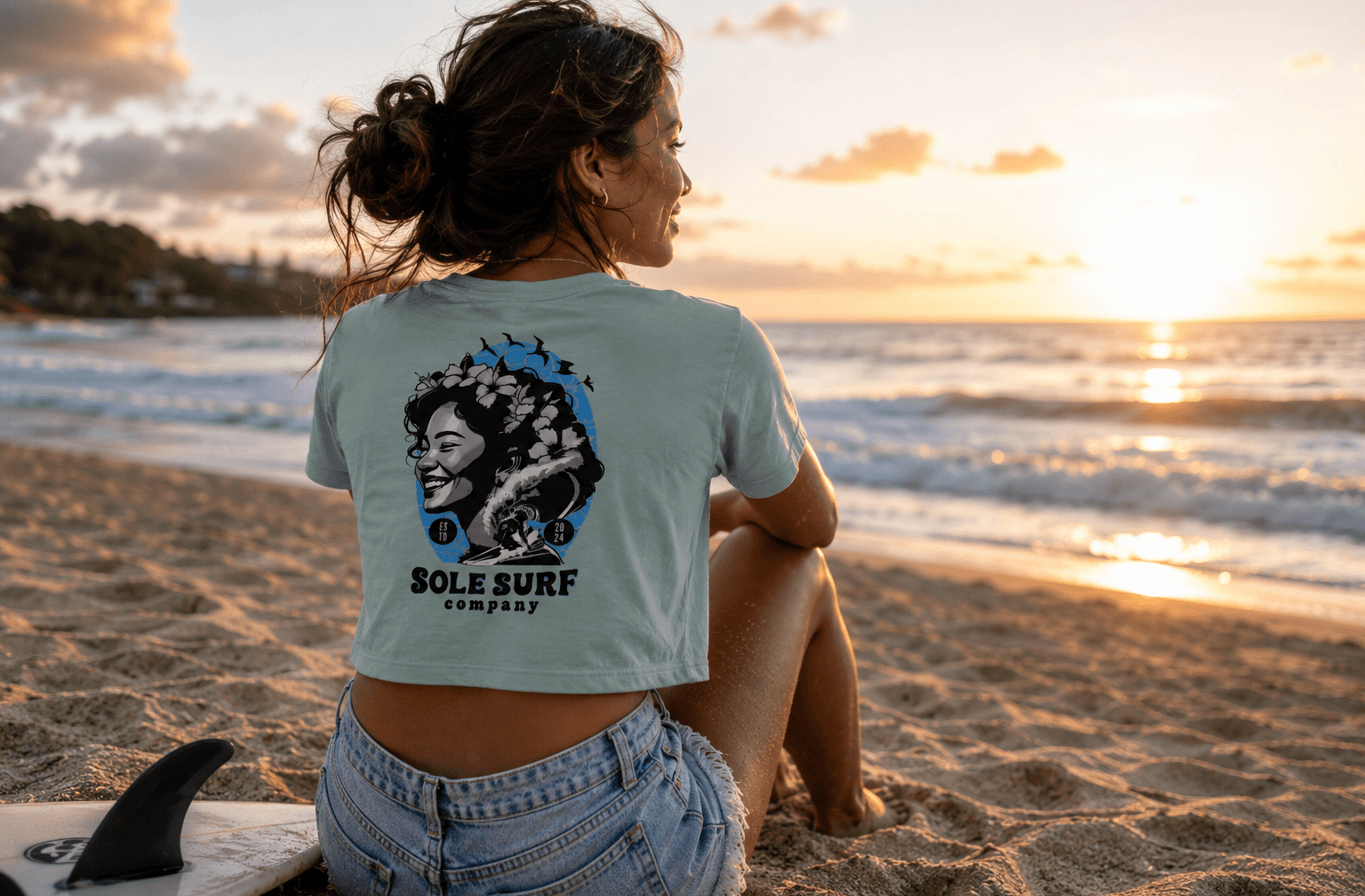 Woman sitting on the beach with a sunset, wearing a t-shirt with 'Sole Surf Company' design.