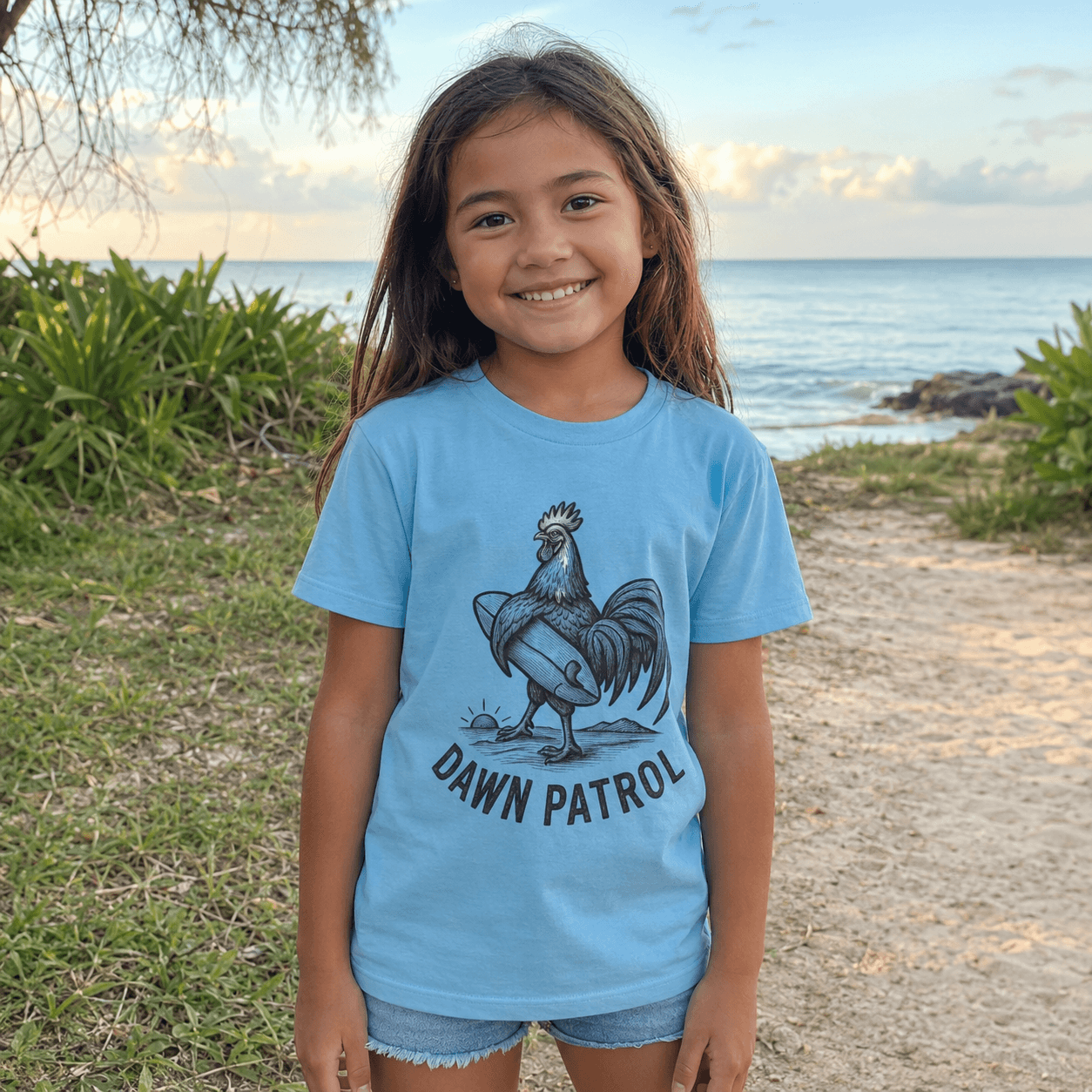 A young girl smiling outdoors on a beach path, wearing a light blue t-shirt with a black graphic of a rooster and the text 'DAWN PATROL'.