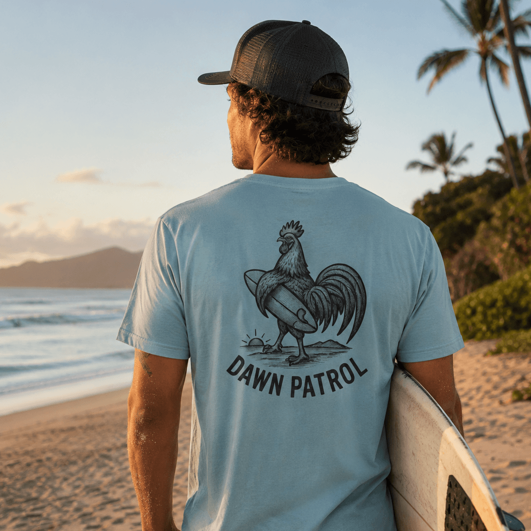 Man on a beach holding a surfboard wearing a t-shirt with a rooster graphic and 'Dawn Patrol' text.