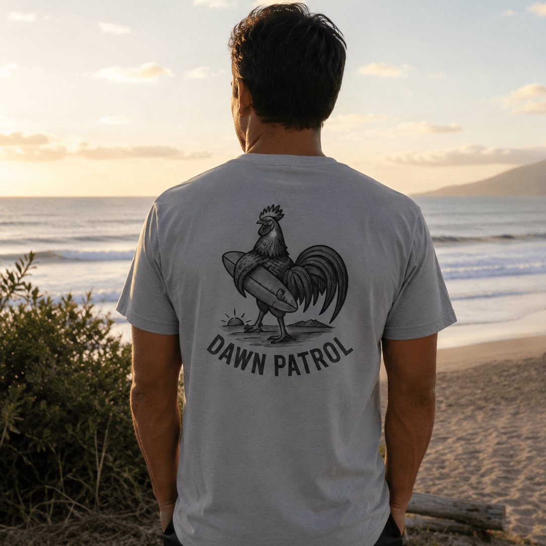Person wearing a t-shirt with a rooster graphic and 'Dawn Patrol' text on a beach at sunset.