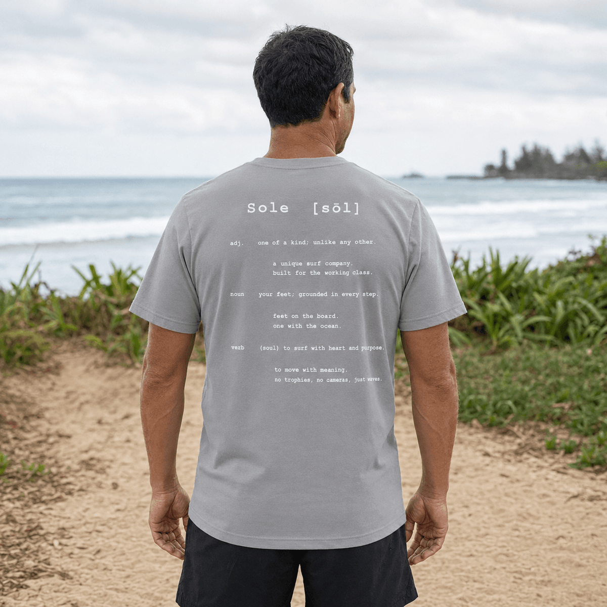 A man standing on a sandy beach with the ocean and greenery in the background, viewed from behind. He is wearing a light gray t-shirt with white printed text on the back reading 'Sole [sûl]' and black shorts.