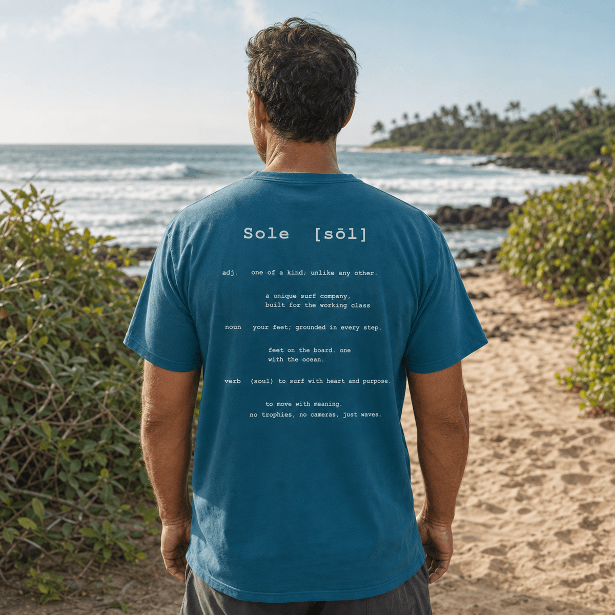A man stands on a sandy beach with the ocean and greenery in the background, viewed from behind. He is wearing a teal blue t-shirt with white printed text on the back reading 'Sole [sɔːl]'.
