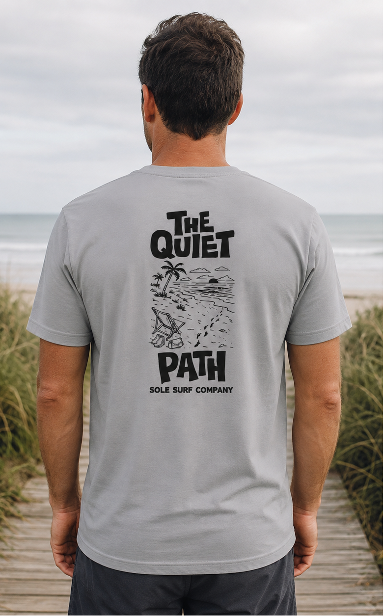 A man stands on a wooden boardwalk at the beach wearing a light gray t-shirt with a black graphic print on the back that reads 'THE QUIET PATH SOLE SURF COMPANY'. A white surfboard rests on the grass to his right, with the ocean and cloudy sky in the background.