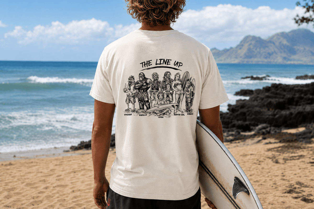 A man standing on a beach, viewed from behind, wearing a cream-colored t-shirt with a black graphic print of surfers and the text 'THE LINE UP'. He is holding a white surfboard against his side, with the ocean, mountains, and sandy beach in the background.
