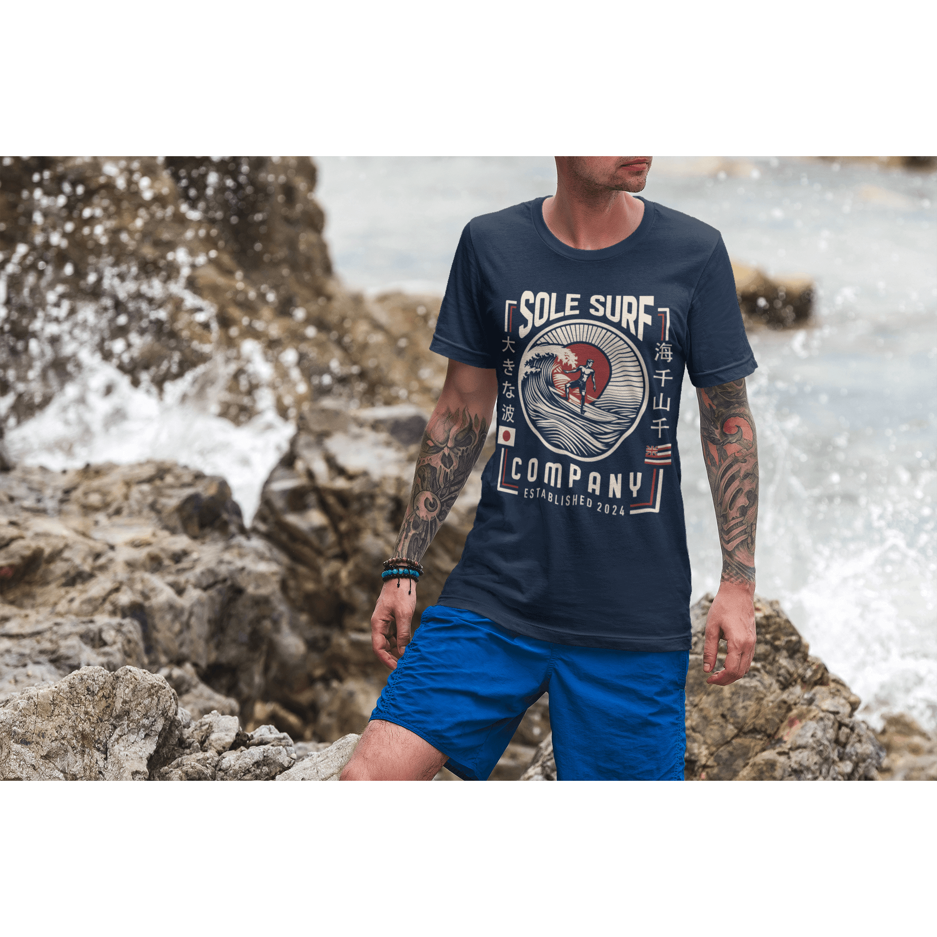 Man wearing a navy Japanese Hawaiian Flag T-shirt on rocky beach, showcasing a relaxed surf style.