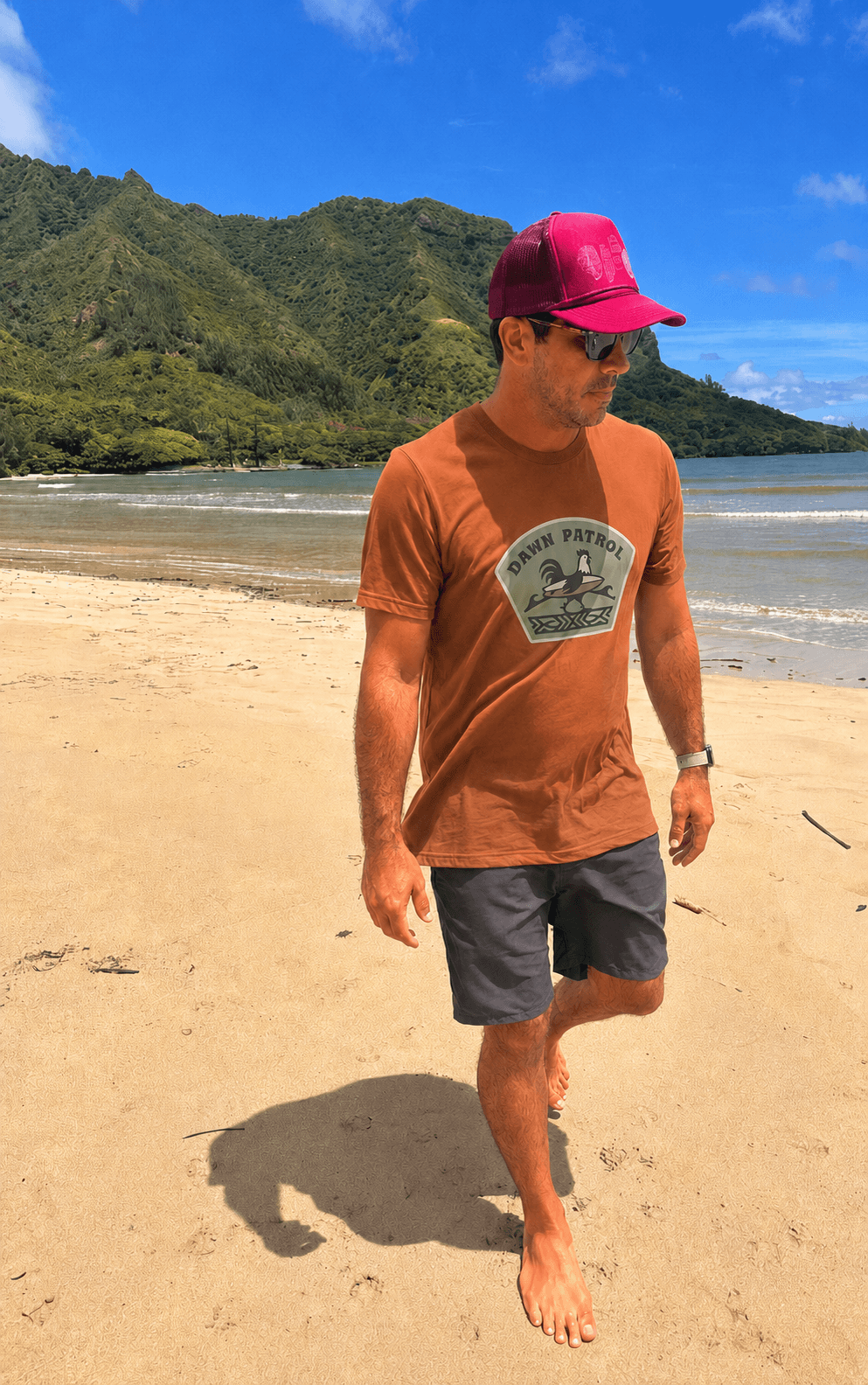 A man walking on a sandy beach wearing a rust-brown t-shirt with a circular graphic that reads 'ORION PATRIOT', dark gray shorts, and a bright pink cap, with ocean and green mountains in the background.