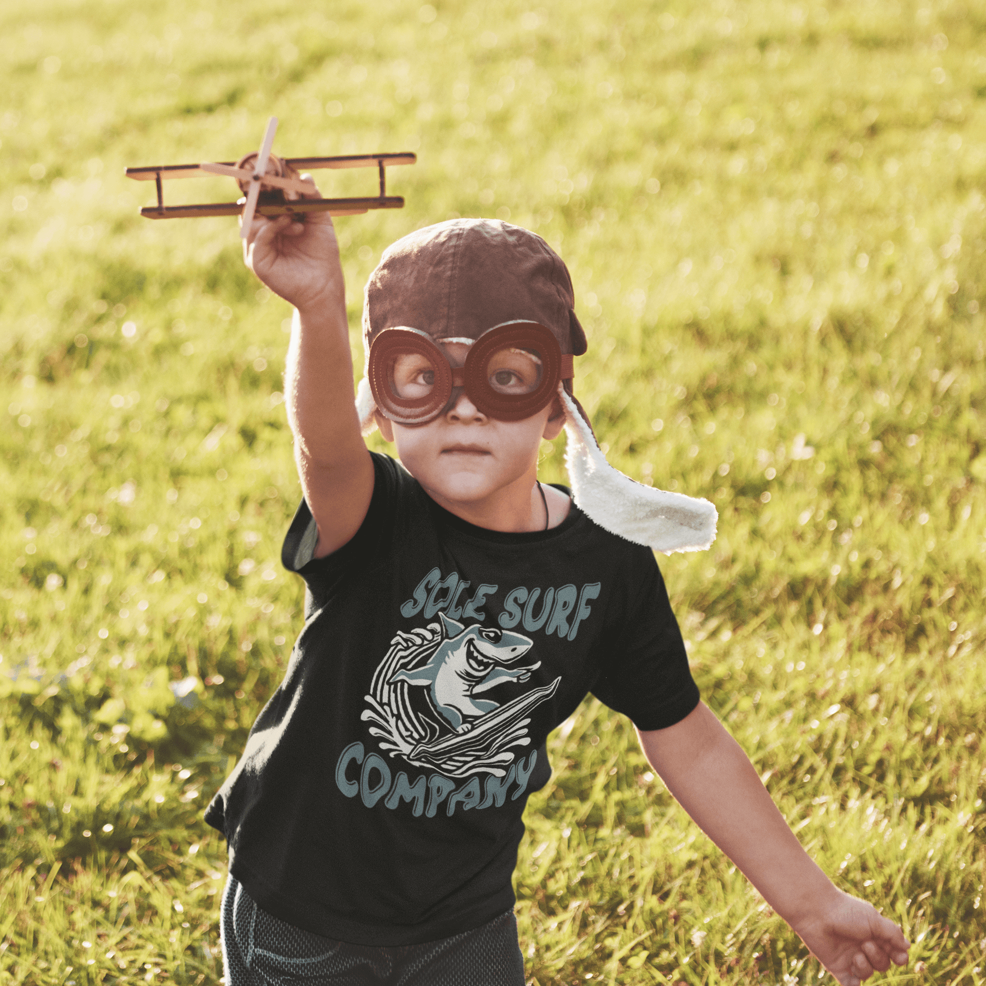 Young child wearing Kids Surfing Shark tee, aviator goggles, and holding a toy plane, playing outdoors.