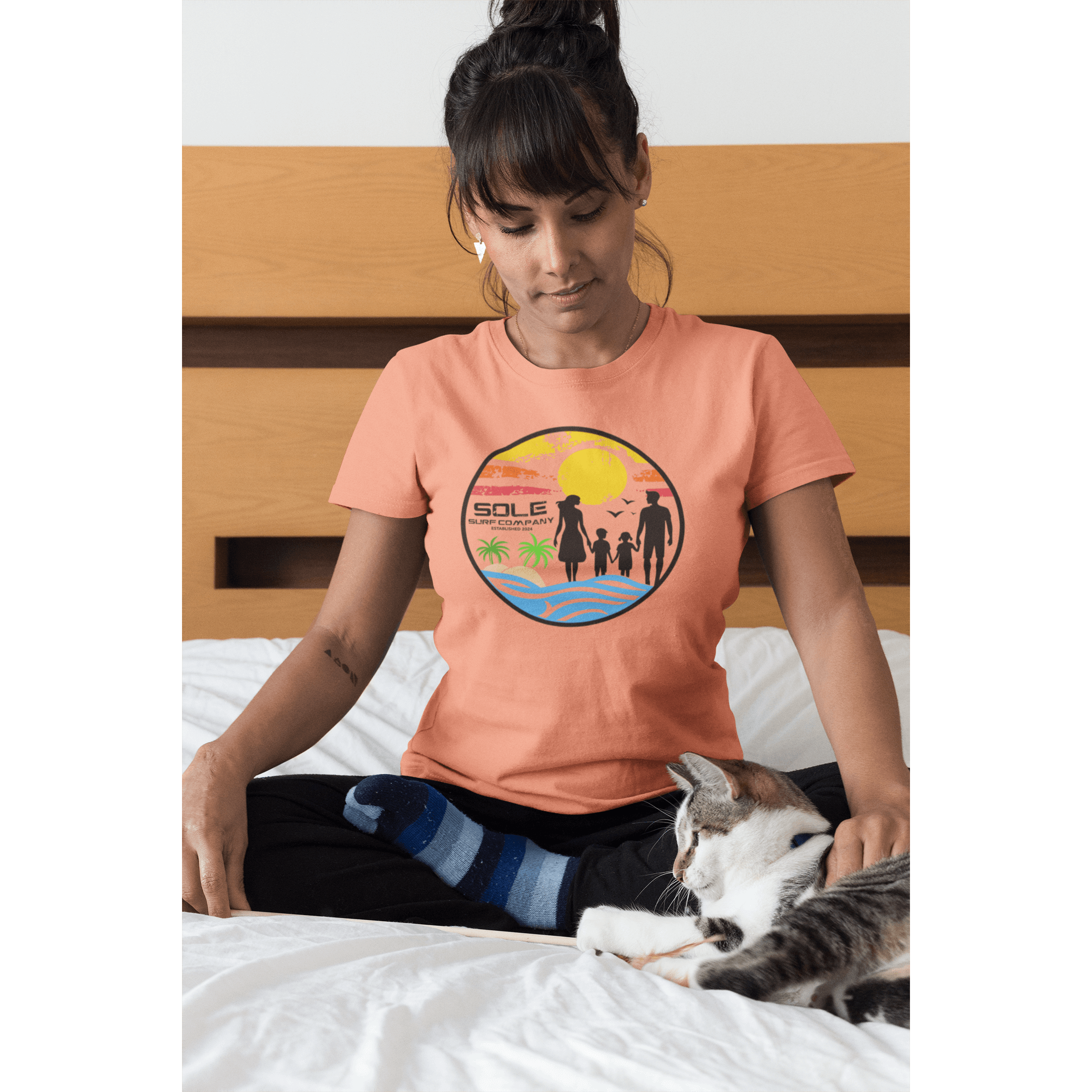 Woman in a peach surf-themed tee sitting on a bed with a cat, embodying a relaxed beach vibe.
